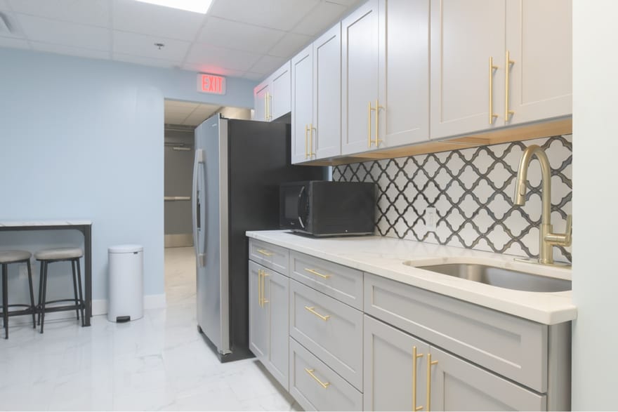 The shared break room: gray shaker cabinetry with brass handles, a patterned backsplash, a full-height fridge, a microwave and a small table with stools.