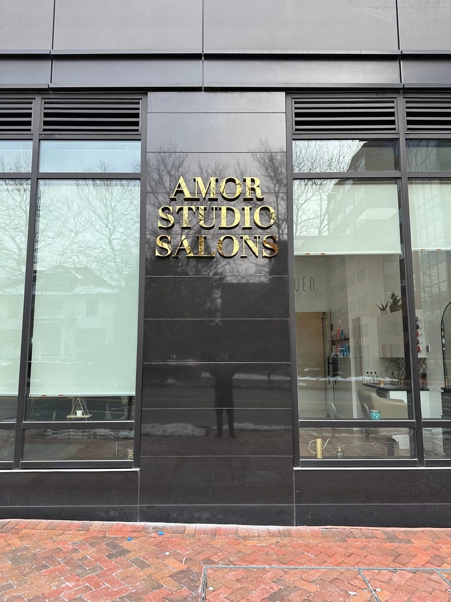 The Amor Studio Salons name in gold leaf, set in three stacked lines of caps on a polished black granite panel at the Bethesda building.