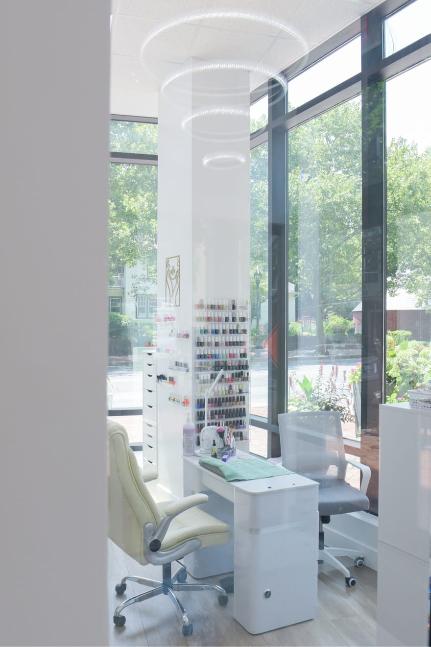 A nail suite seen through its glass door: a manicure table, a wall rack of polish, a client chair and a ring light, in front of full-height windows.