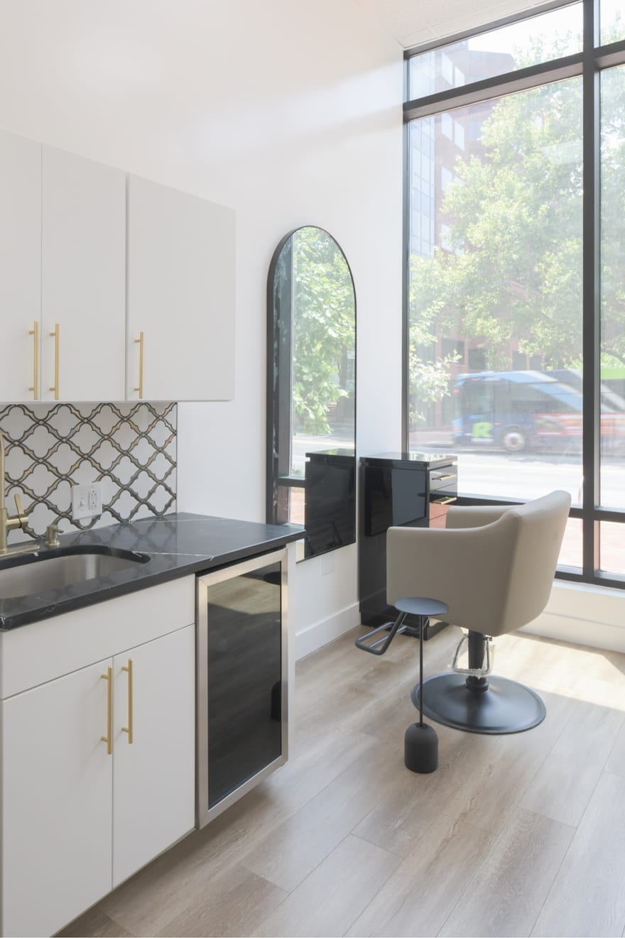 A fitted salon suite: white cabinetry with brass handles, a black stone counter and shampoo sink, an under-counter beverage fridge, an arched black mirror and a styling chair beside a floor-to-ceiling window onto the street.