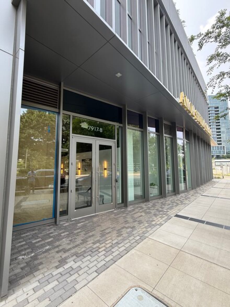 The Westpark Drive frontage at 7917-B: a gray glazed storefront under a deep colonnade, with the gold Amor lettering along the facade and a Tysons tower rising behind.