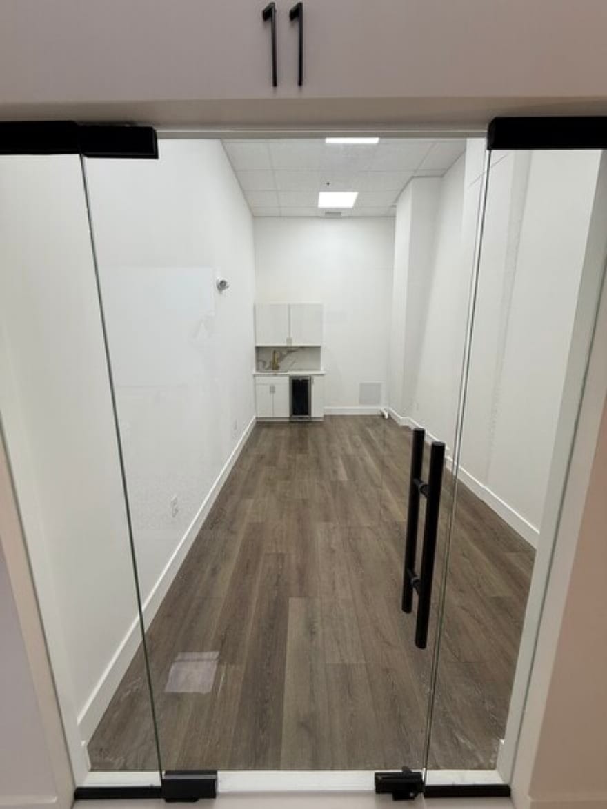 An empty suite at the Tysons building seen through its open full-height glass doors: wood-look flooring, white walls, and a white kitchenette with a sink at the far end.